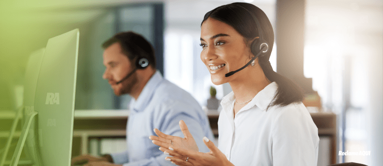 Mulher sorrindo olhando para tela do computador. Em segundo plano um home de camisa azul.